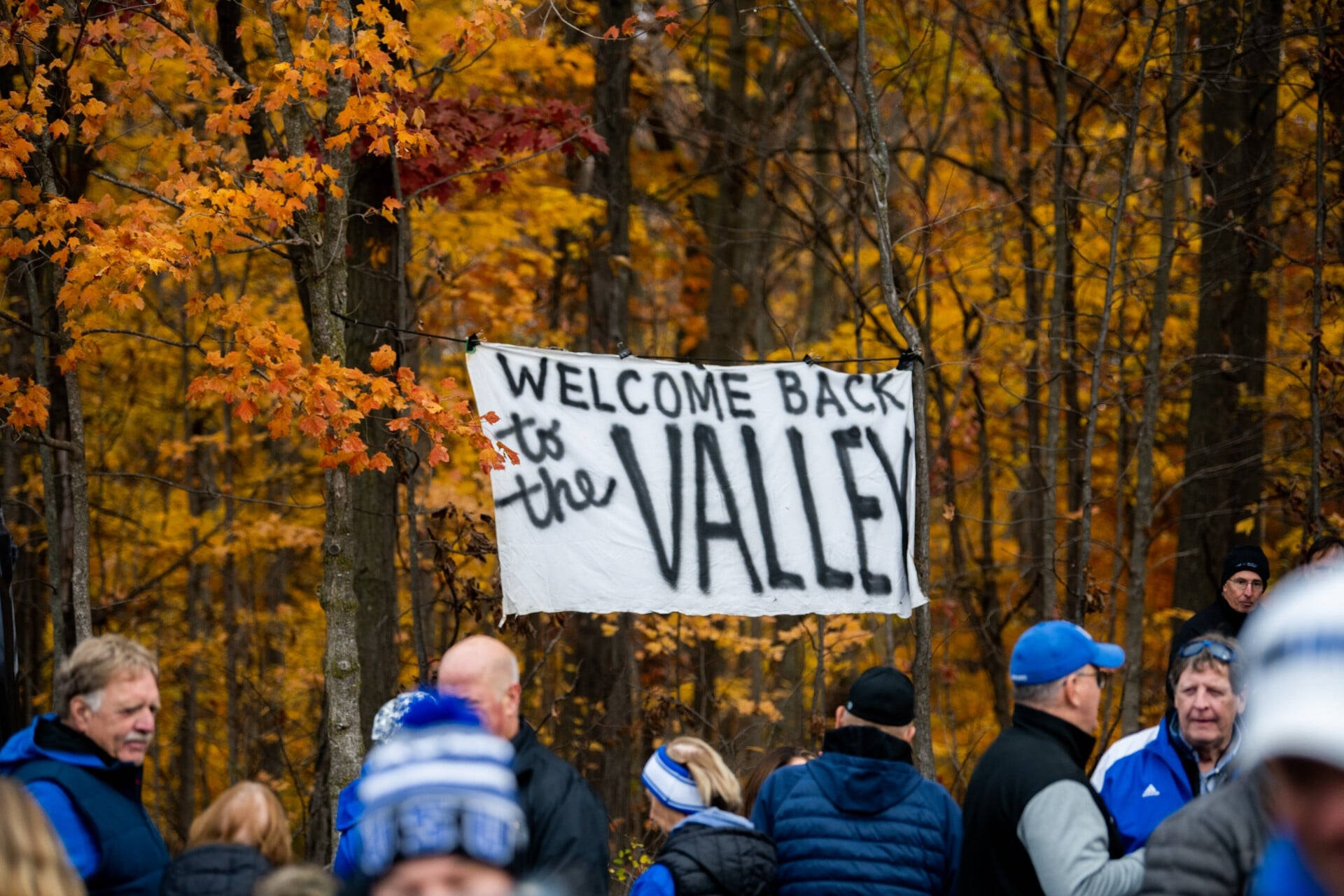Sign reads Welcome Back to the Valley, colorful fall trees in background, people gathering in winter hats and coats